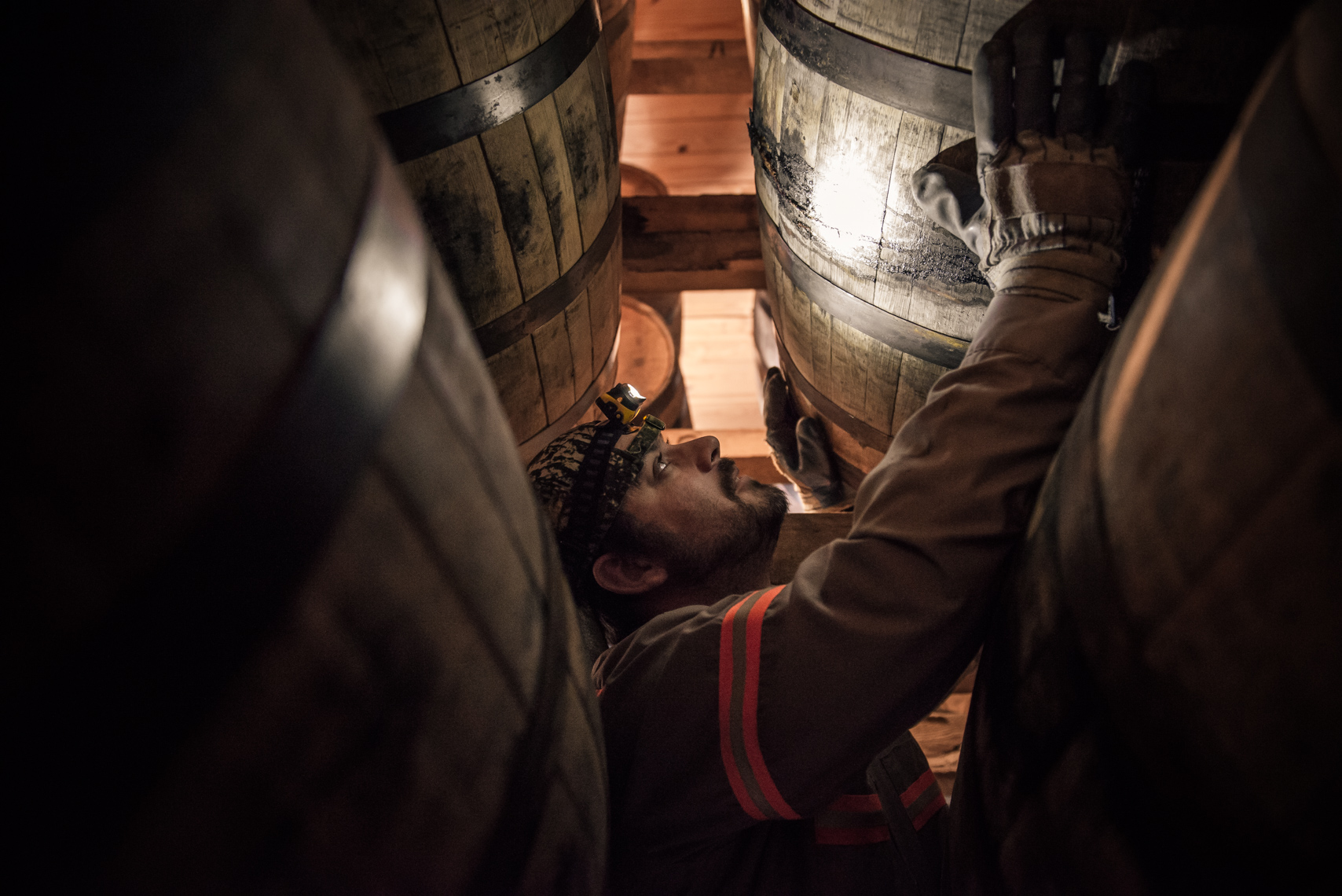 Photo of a man wearing a headlamp climbs on racks in a darkly lit warehouse of aging whiskey barrels |  Kristina Krug Nashville photojournalist.